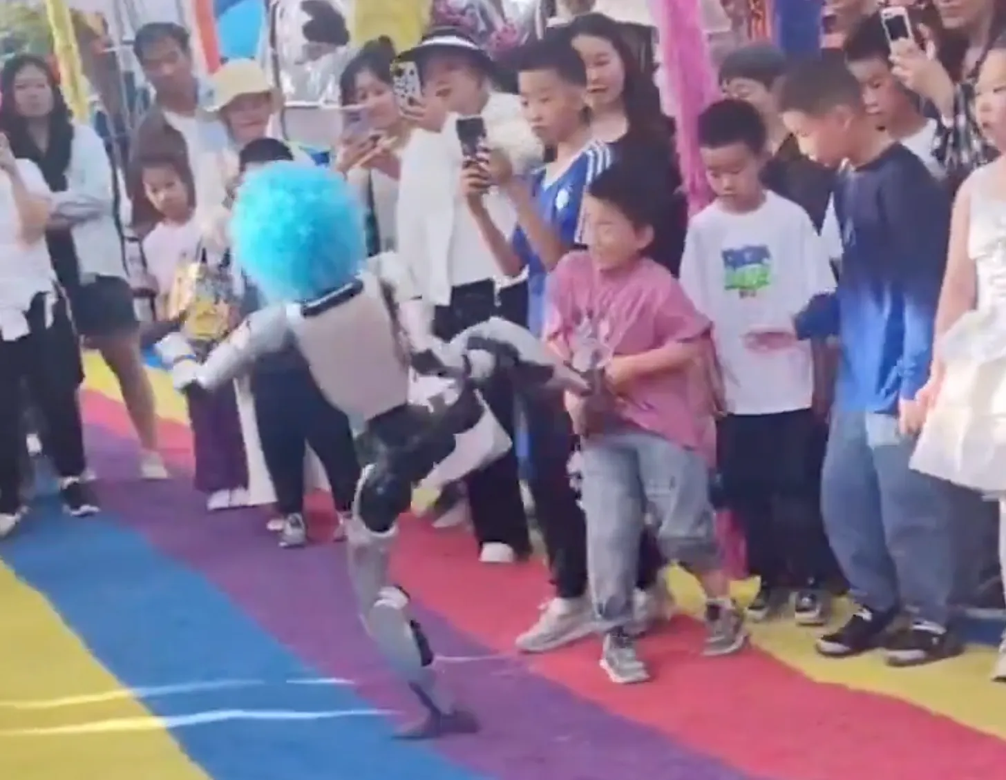 A small humanoid robot in a fluffy turquoise wig kicks a child in the stomach on a rainbow-striped mat, one leg lifted and arms swinging, while a dense crowd of children and adults presses in around it, several holding up phones to film.